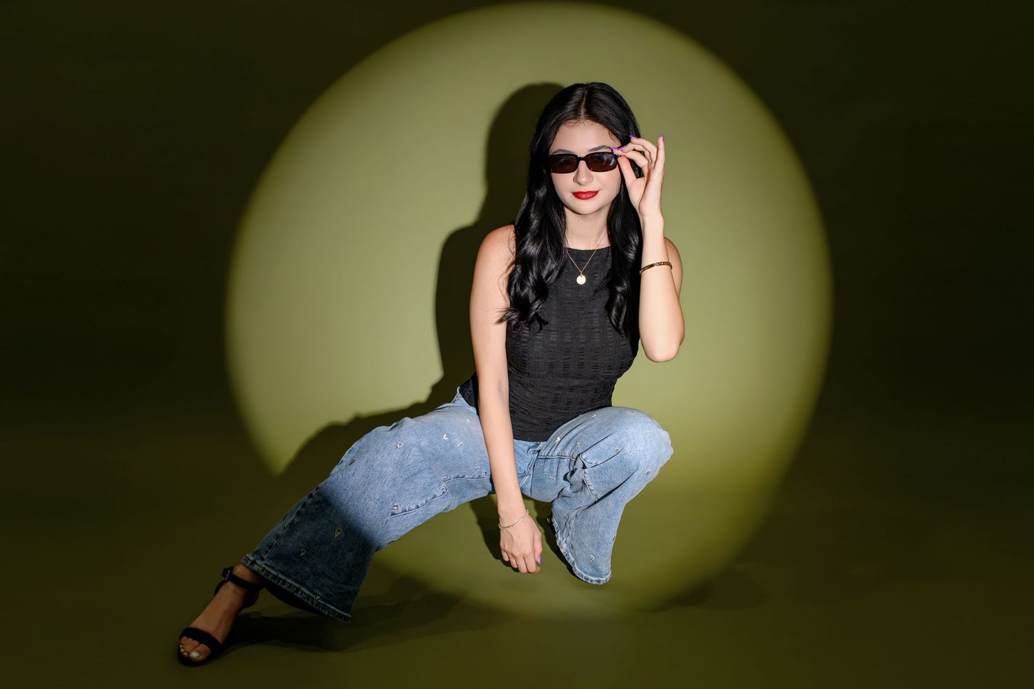 Young woman in black top and wide-leg jeans poses on green background, wearing sunglasses in stylish fashion shoot.