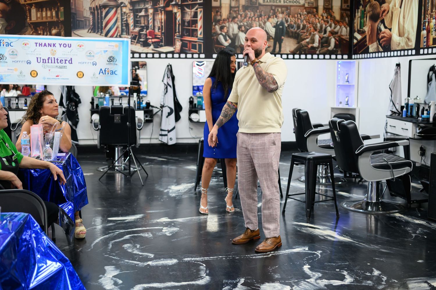 Man speaking in a modern barbershop with women nearby, thank you sponsors banner visible in background.