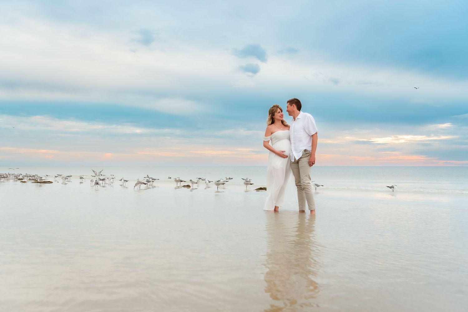 Couple in white standing in shallow ocean water at sunset with seagulls on a serene beach.