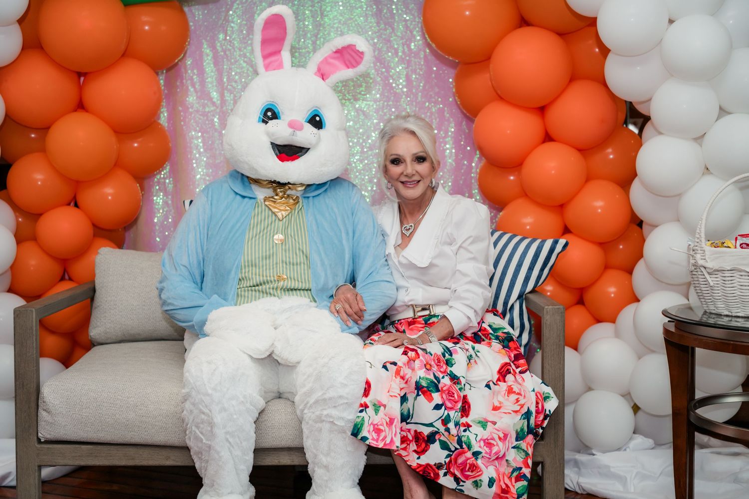 Woman posing with Easter Bunny character on a sofa surrounded by orange and white balloon arch decorations.
