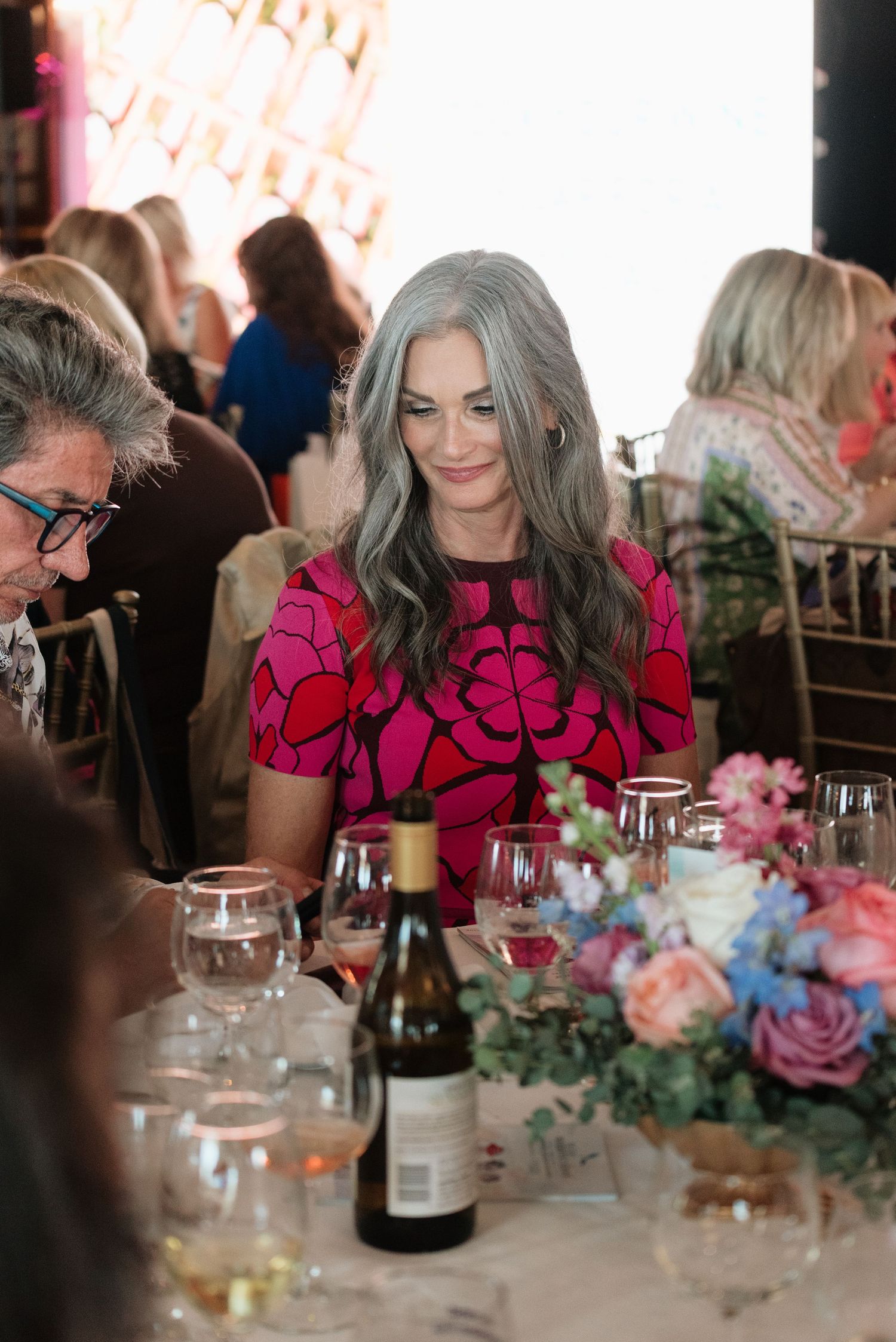 A woman in a vibrant pink floral dress sits at an elegant dinner table with wine glasses and floral centerpieces.
