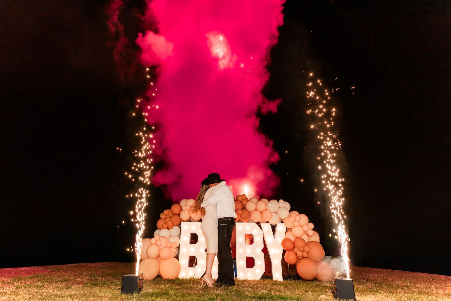 Pink smoke and sparklers illuminate a large glowing 'DADDY' sign during an outdoor nighttime gender reveal celebration.