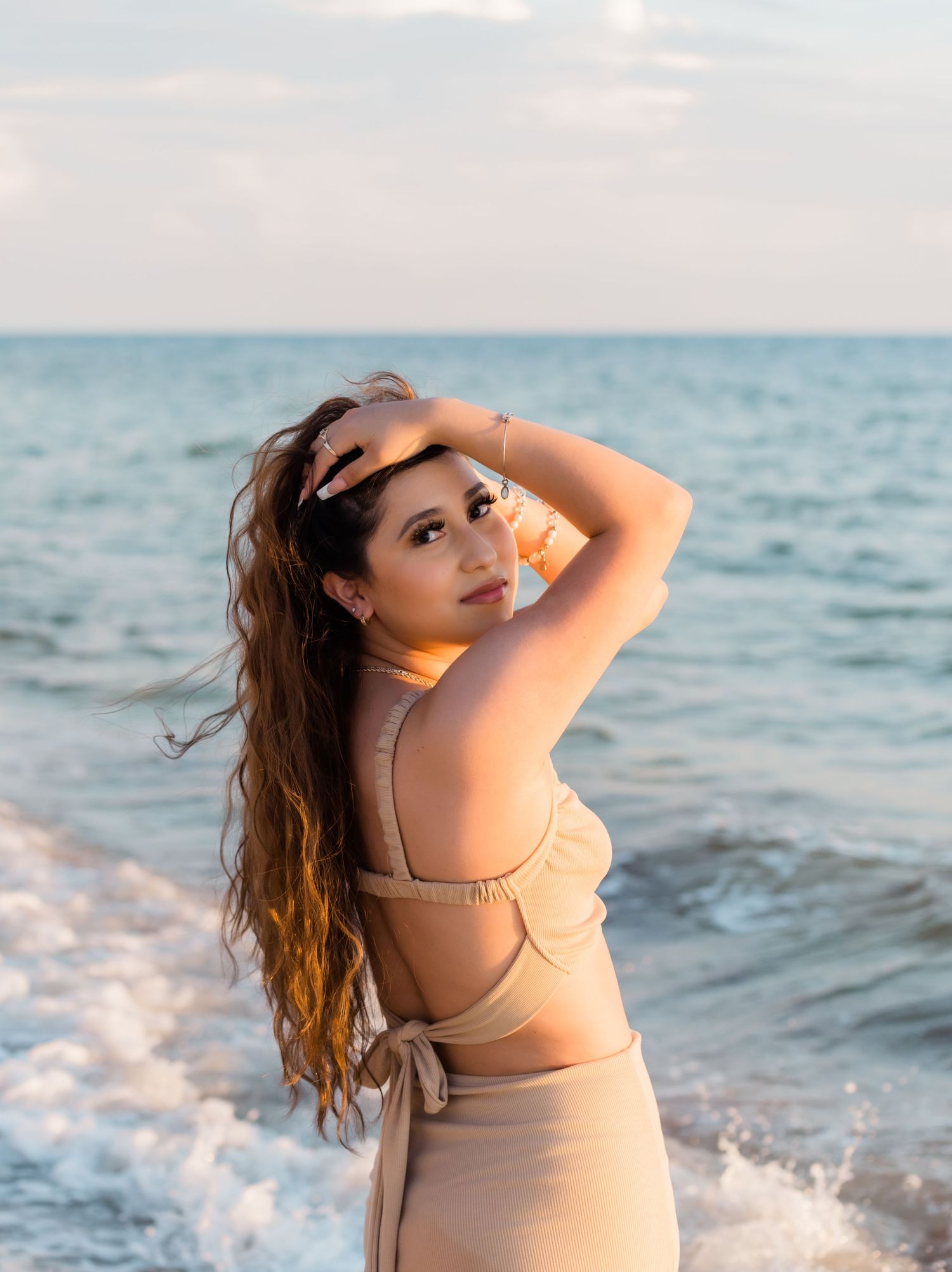Woman in beige swimwear poses on beach at sunset with ocean waves and wind-blown hair.