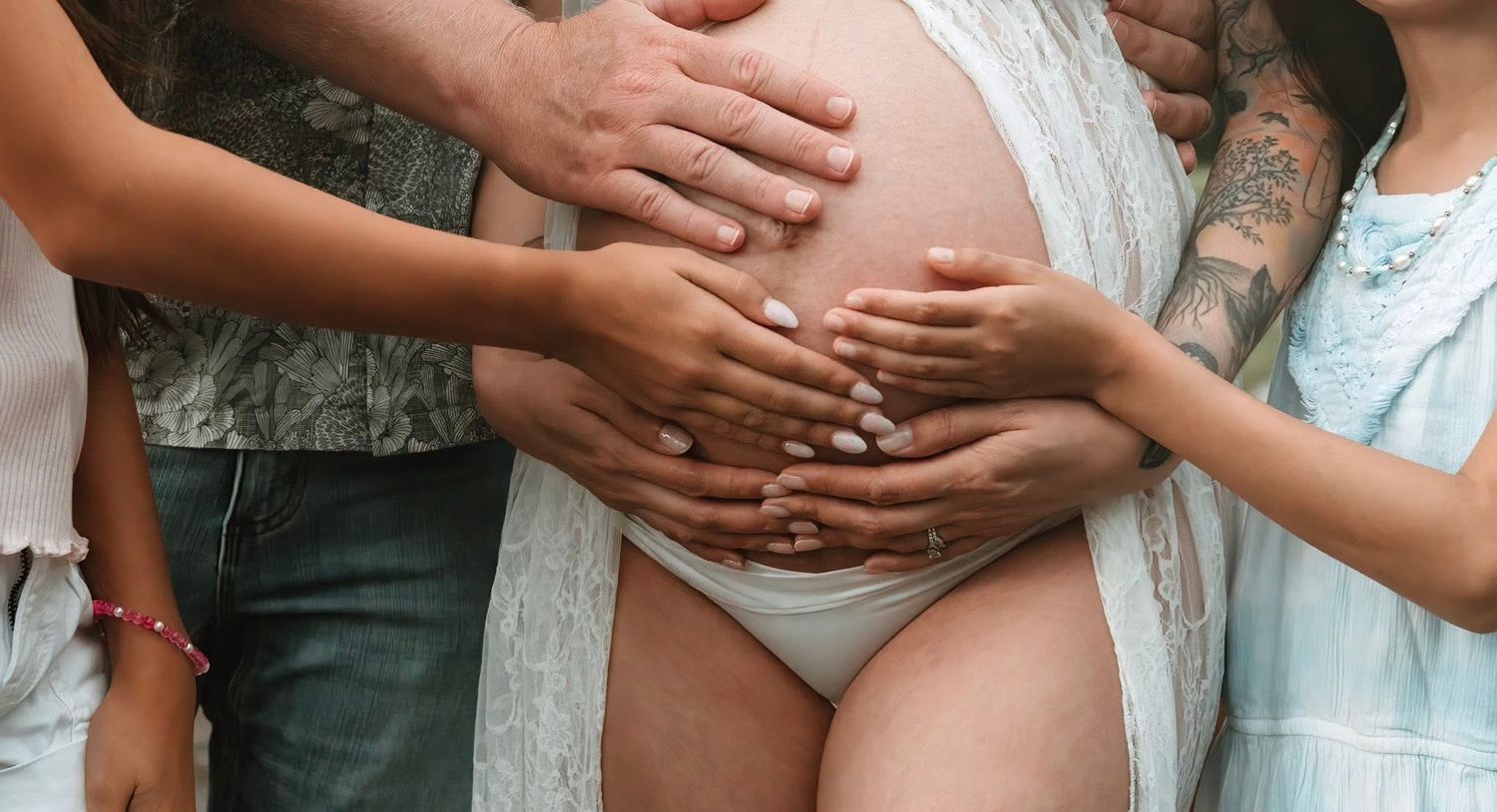 Family members placing hands together on a pregnant belly wearing white lace against a soft neutral background.