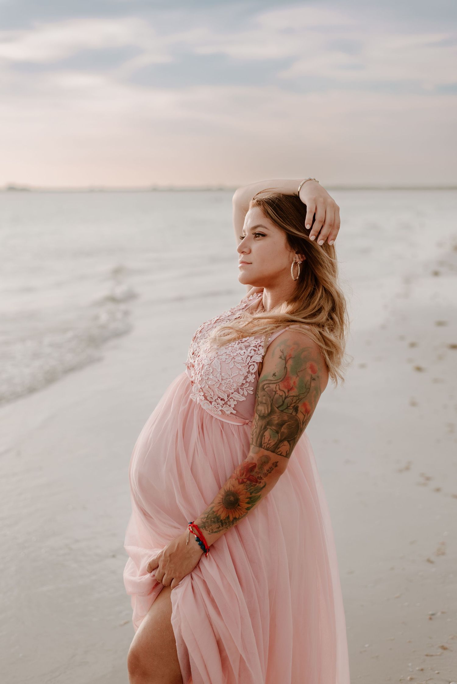 A maternity photo session on a beach features a pregnant woman in a flowing pink dress against a cloudy sky.