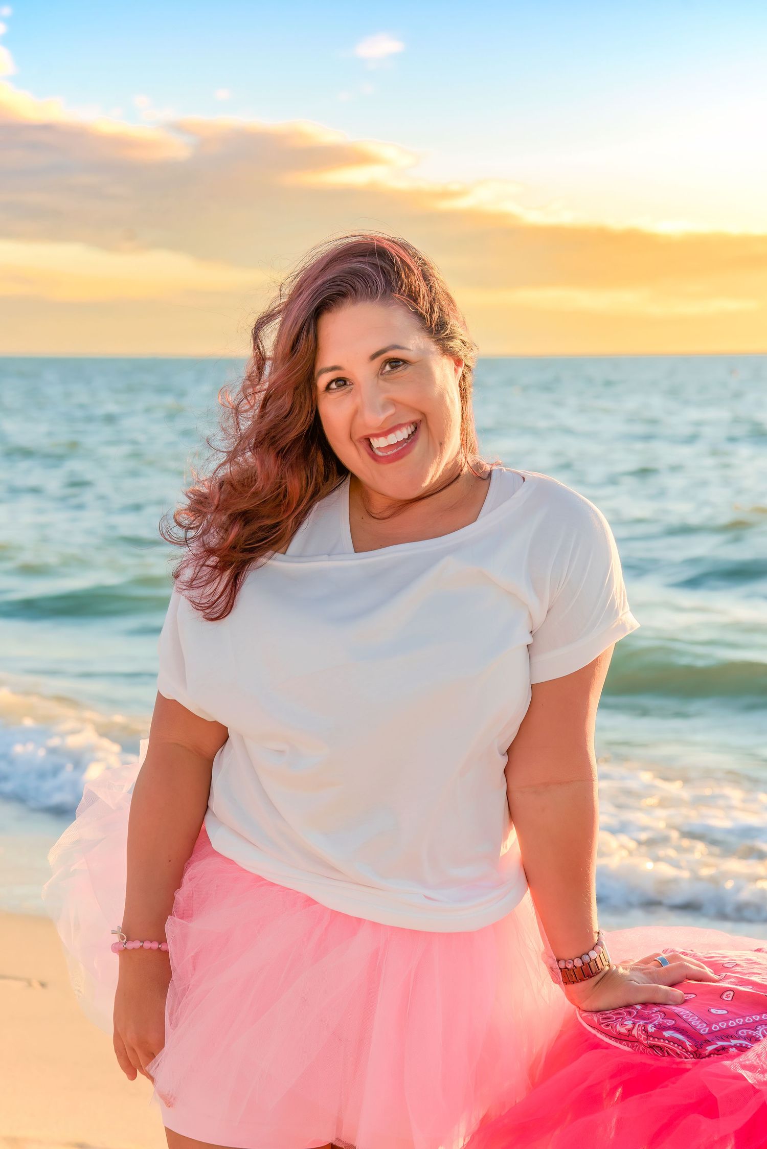 Person in white t-shirt and pink tulle skirt poses joyfully on beach at sunset with ocean waves in background.