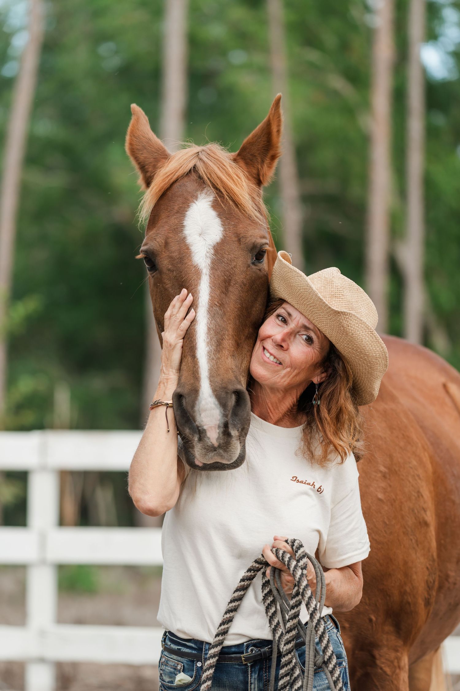 A smiling woman in a cowboy hat affectionately holds a chestnut horse with a white blaze near a white fence.