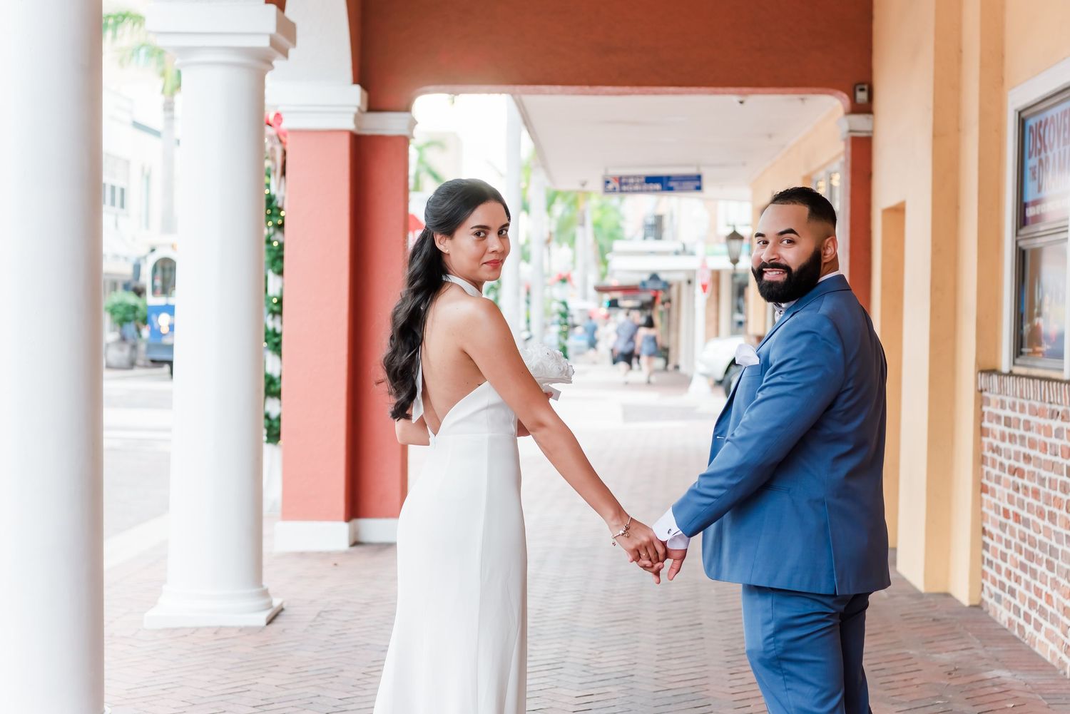 A couple in wedding attire holds hands while standing in a covered outdoor walkway with coral pink columns.