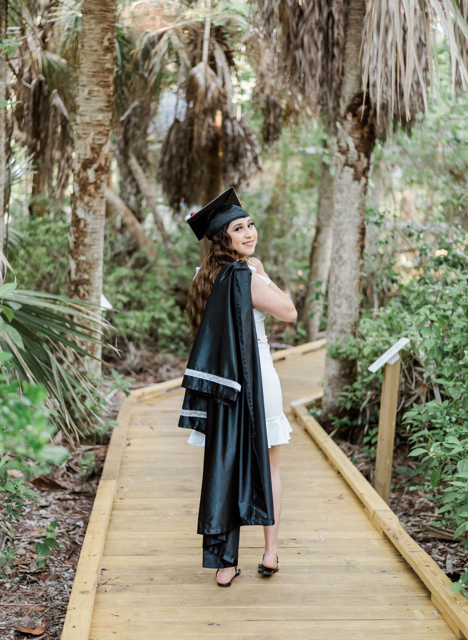 Graduate in black cap and gown standing on a wooden boardwalk path surrounded by tropical palm trees and foliage.