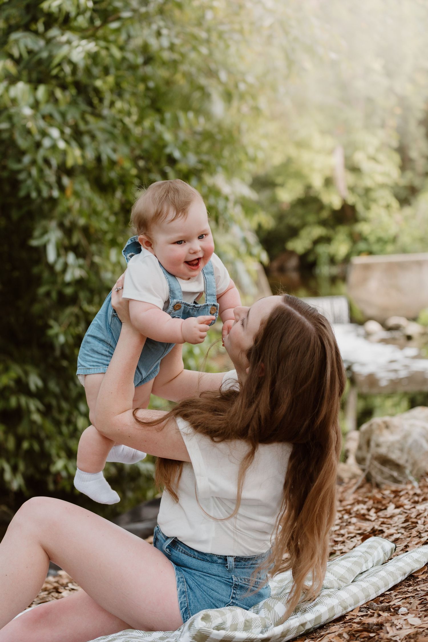 Mother and baby laughing together while playing on a blanket outdoors in a natural garden setting.