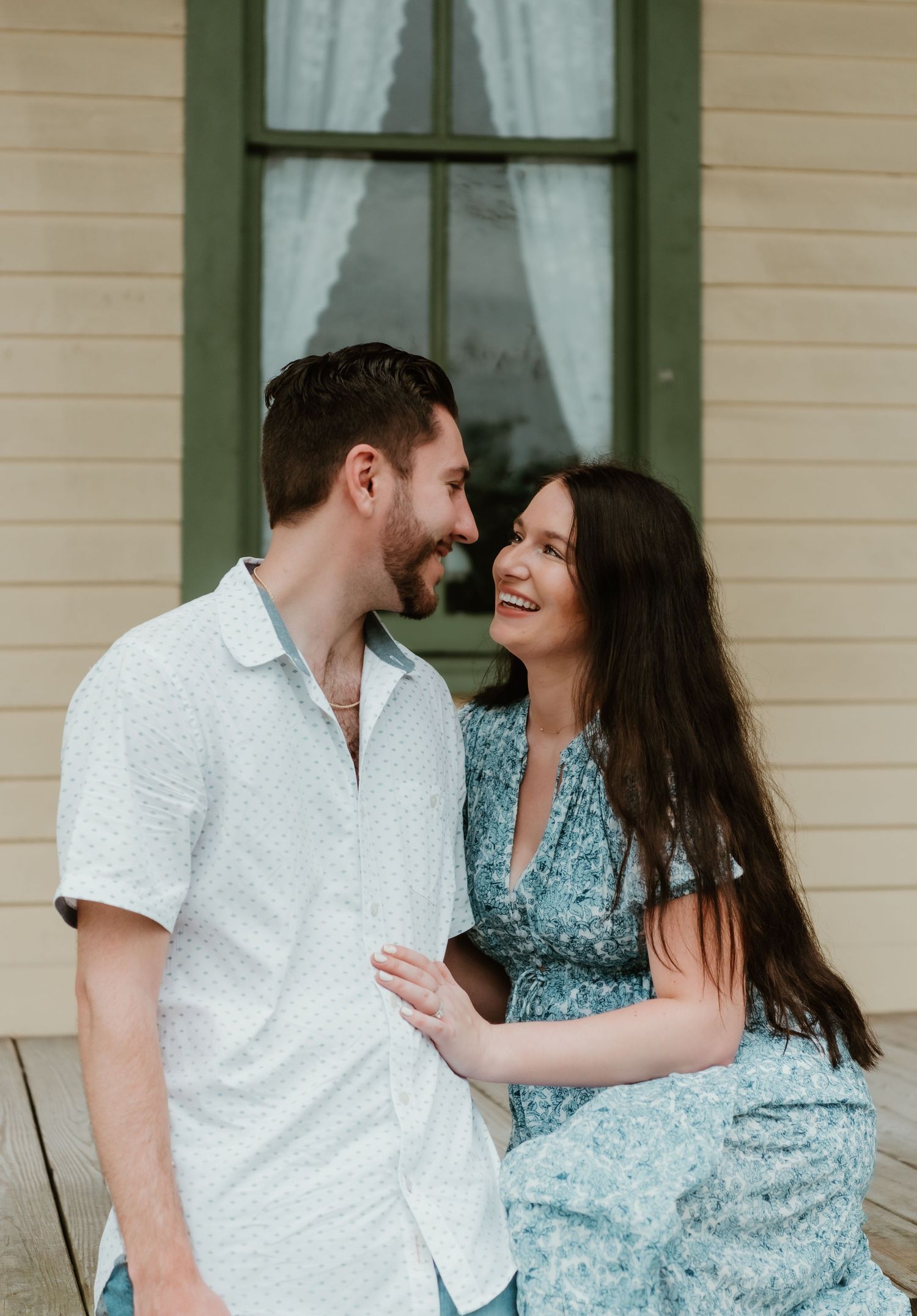 A couple sharing a joyful moment while standing on a porch with white siding and green trim windows.