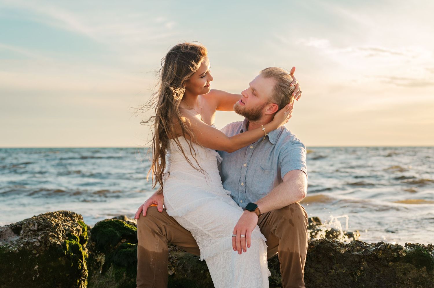 Couple sharing a romantic moment on rocky shoreline at sunset with ocean waves in the background.