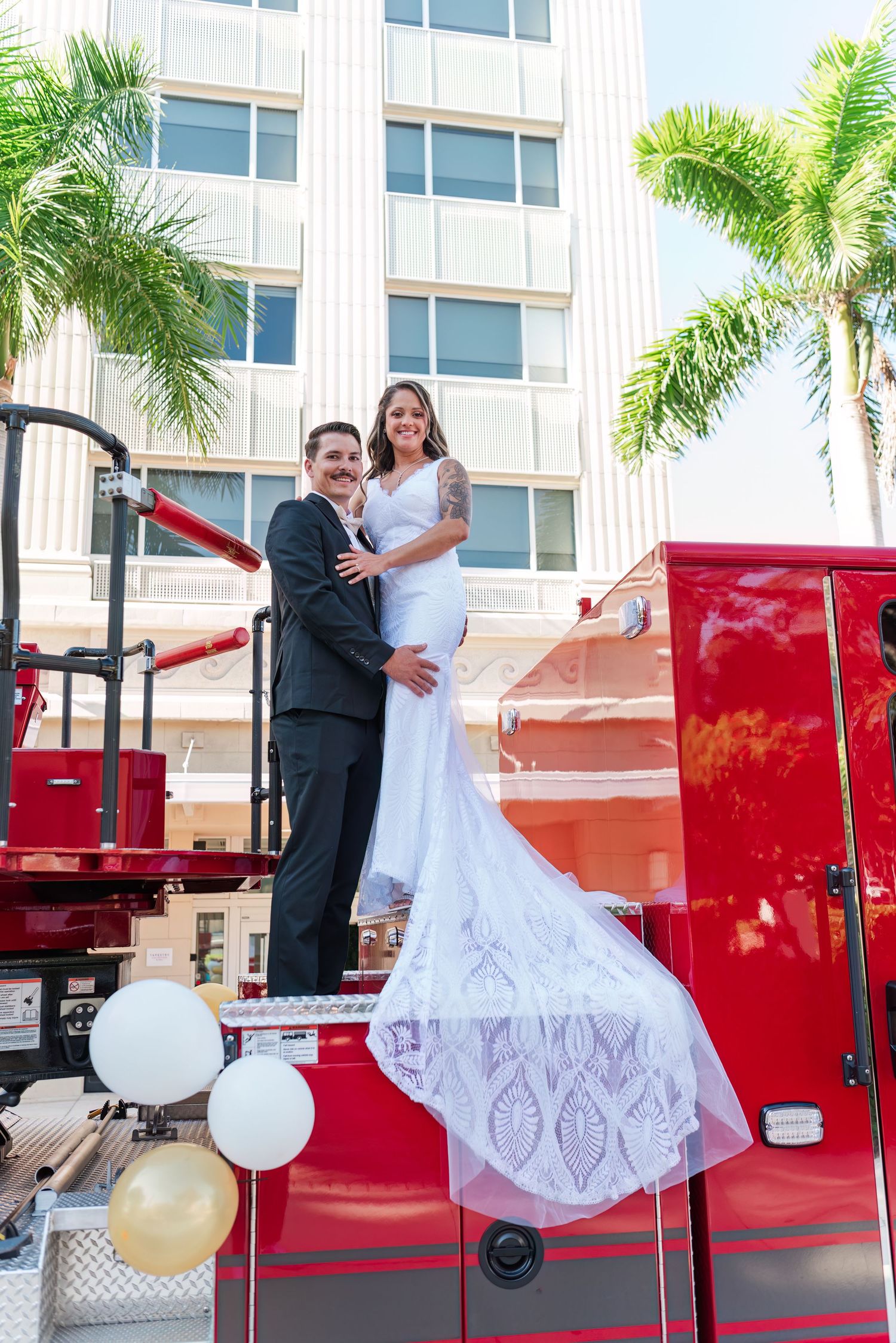 Bride in white gown and groom in black suit stand atop a red fire truck with palm trees in background.