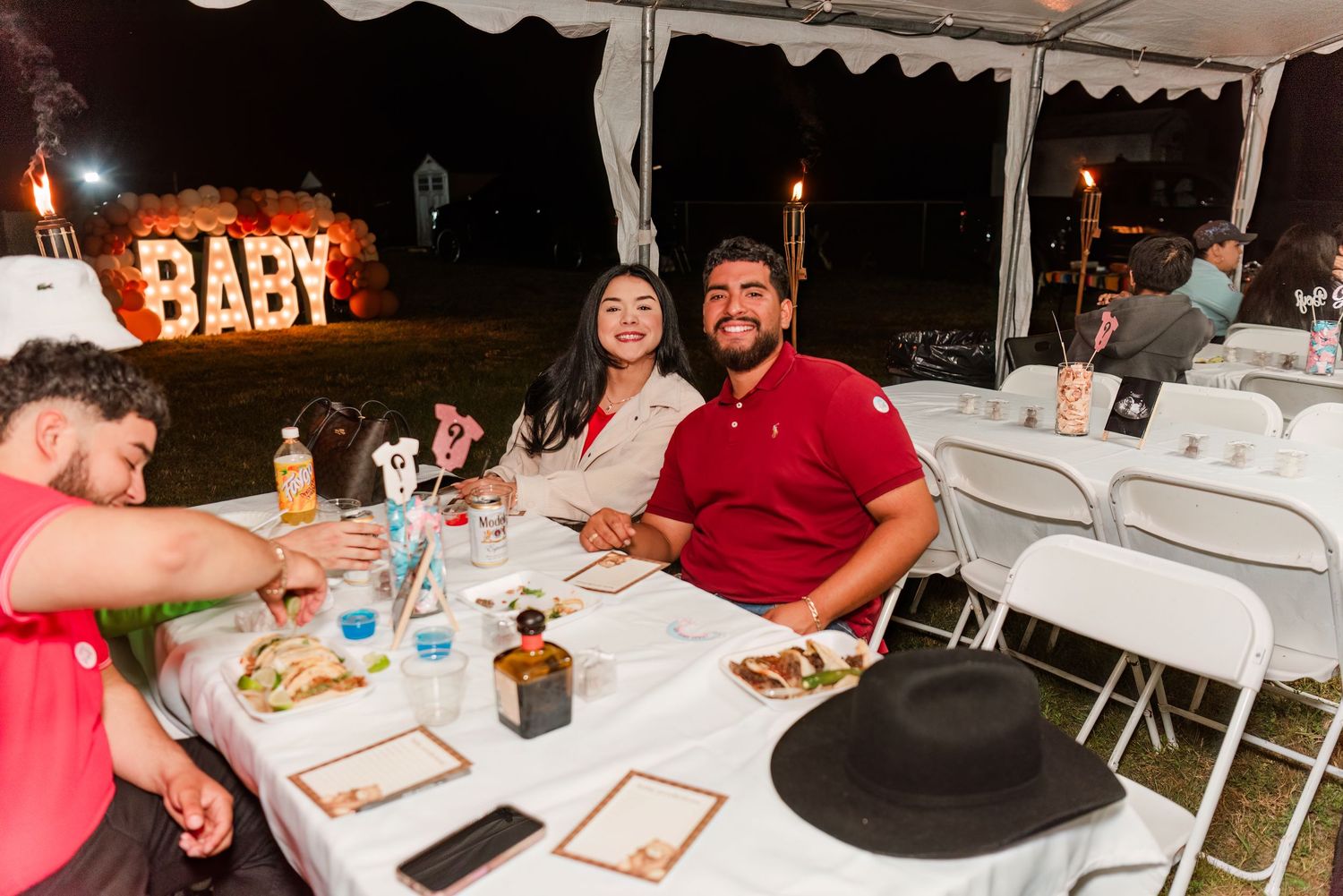 A panoramic view of a casual outdoor evening party with white tables and chairs set up under string lights.