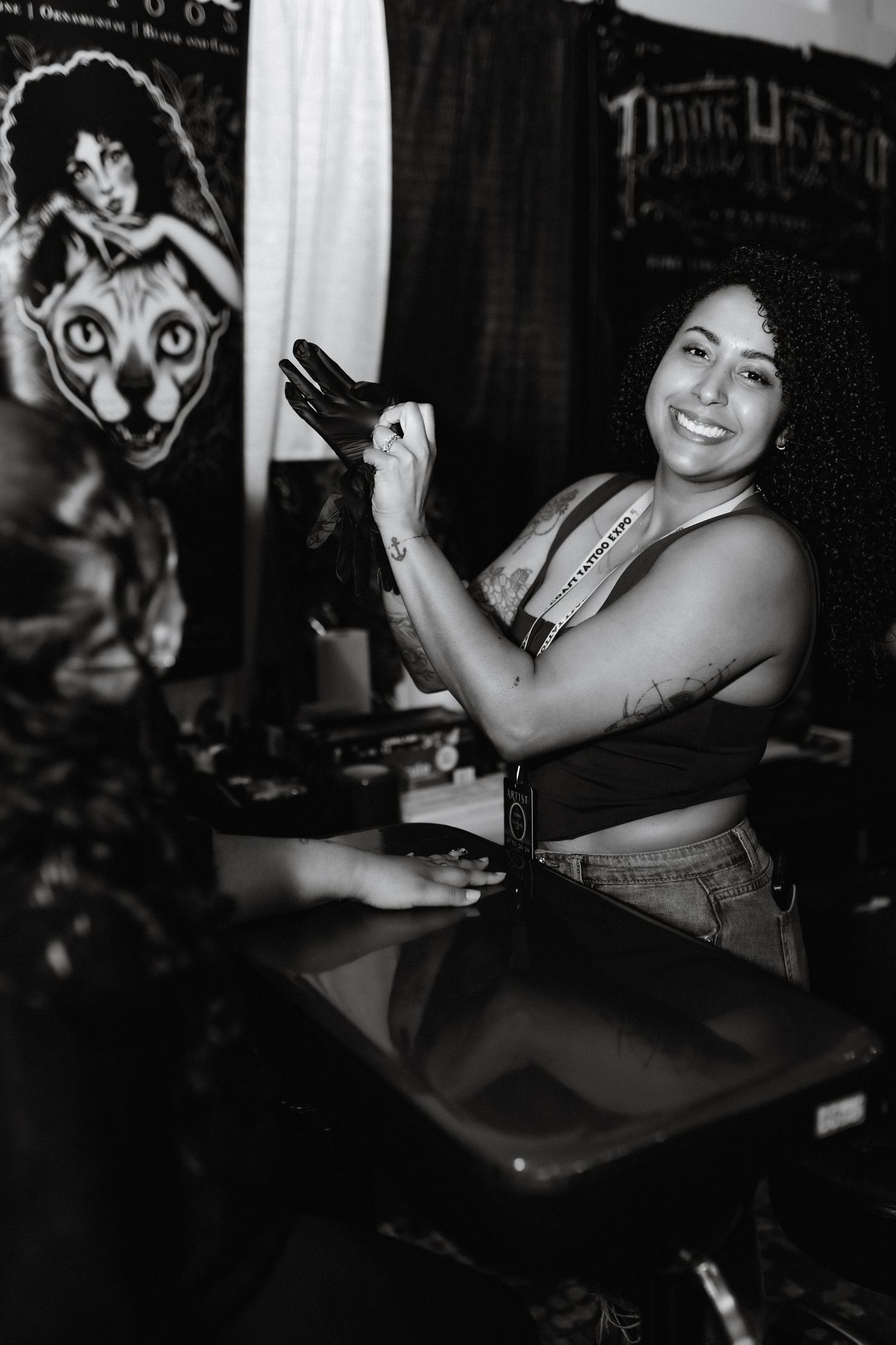 Smiling female tattoo artist in black top putting on gloves at tattoo convention booth in black and white photo.