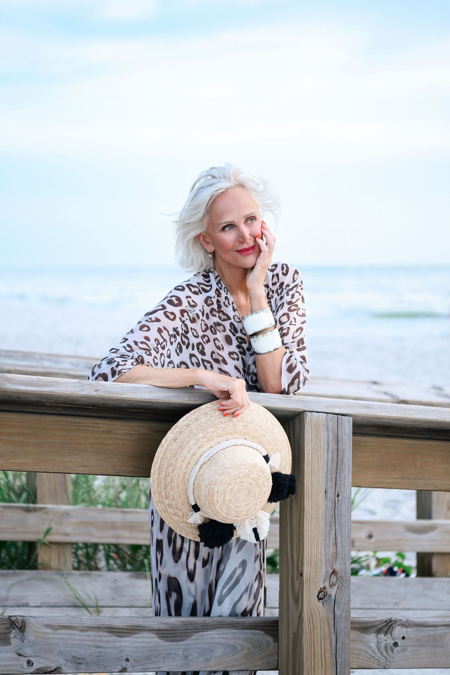 Elegant older woman in leopard print dress holding straw hat, leaning on wooden beach railing with ocean view.