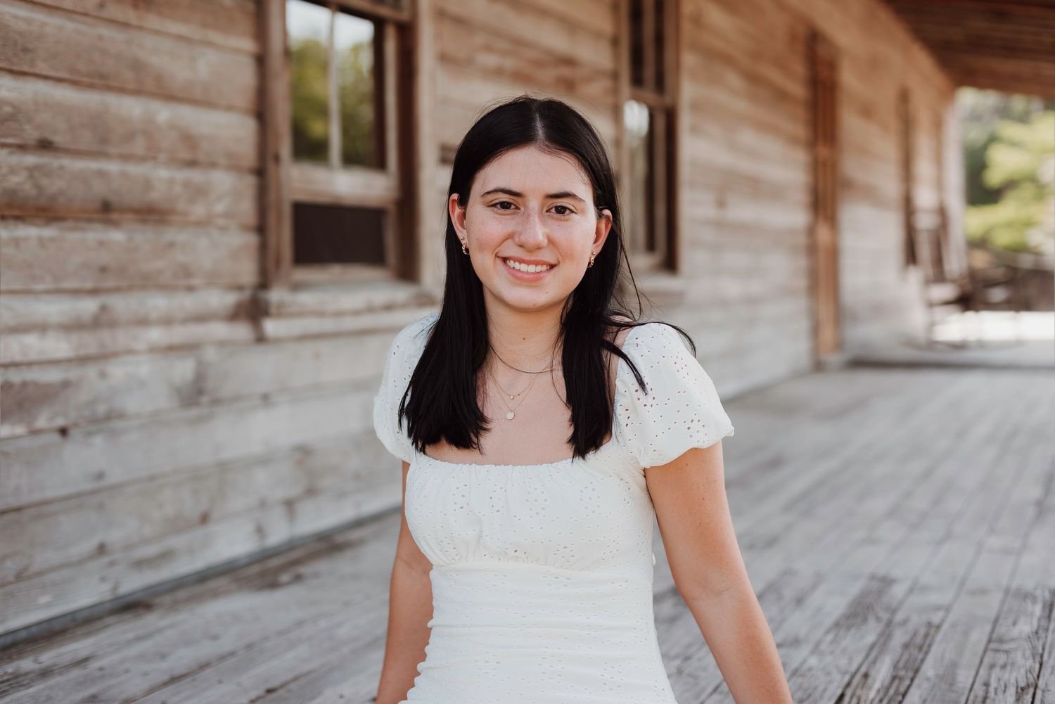 A woman in white dress smiles on a wooden deck during an outdoor photo session.
