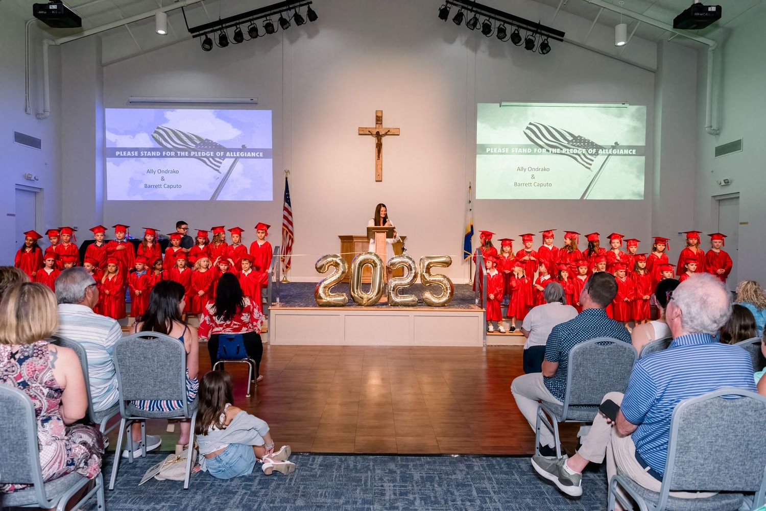 A church auditorium with a cross mounted on the wall and people wearing red seated in rows during a ceremony.