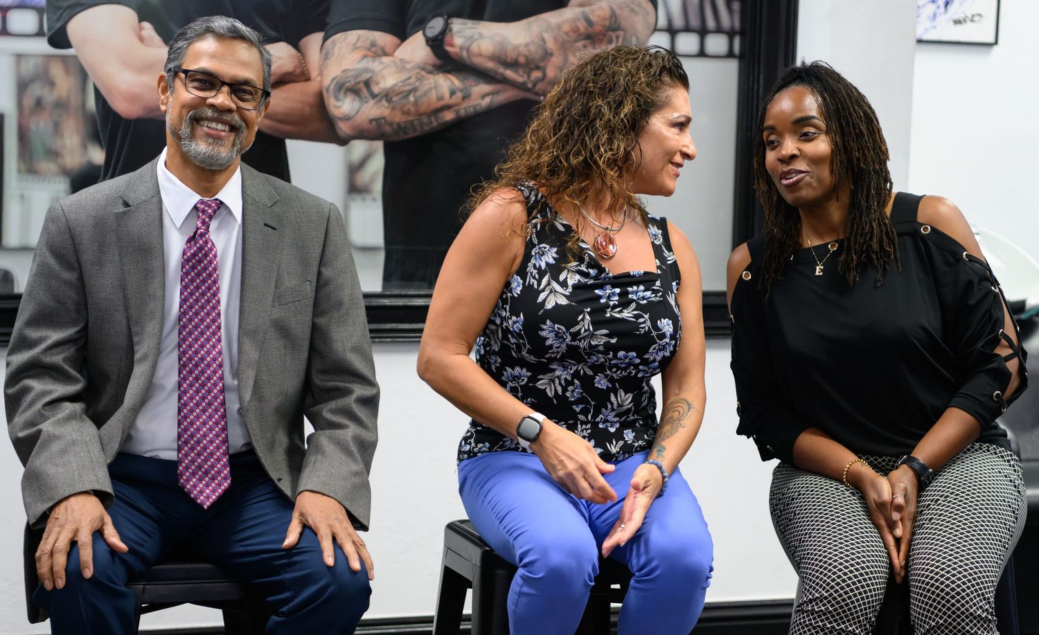 Three people smiling and laughing while seated in a bright room, including a man in a gray suit and two women.
