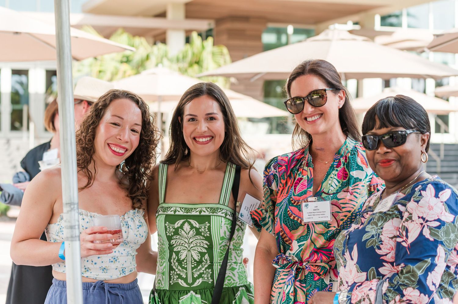 Four women smiling together at an outdoor event on a sunny day.