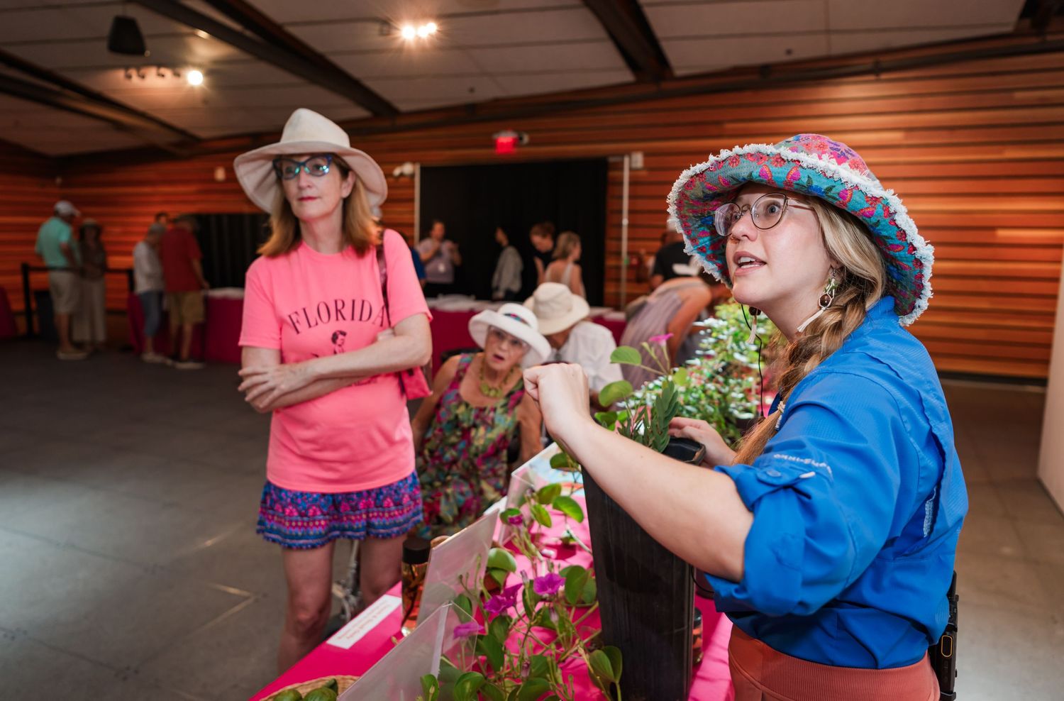 Two girls in colorful hats examine plants at an indoor gardening event in a rustic wooden barn setting.