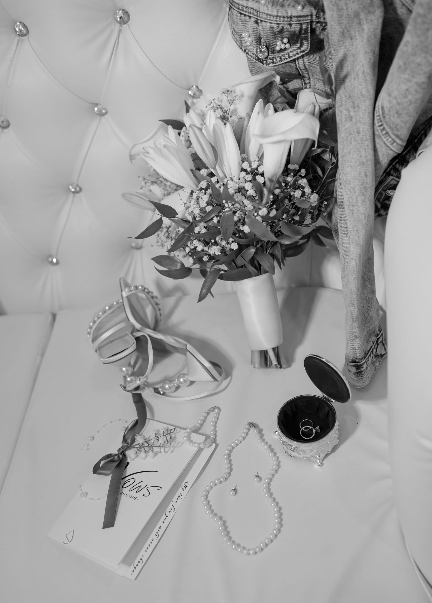 Black and white flat lay of wedding accessories including bouquet, heels, pearl necklace, ring box, and invitation on tufted surface.