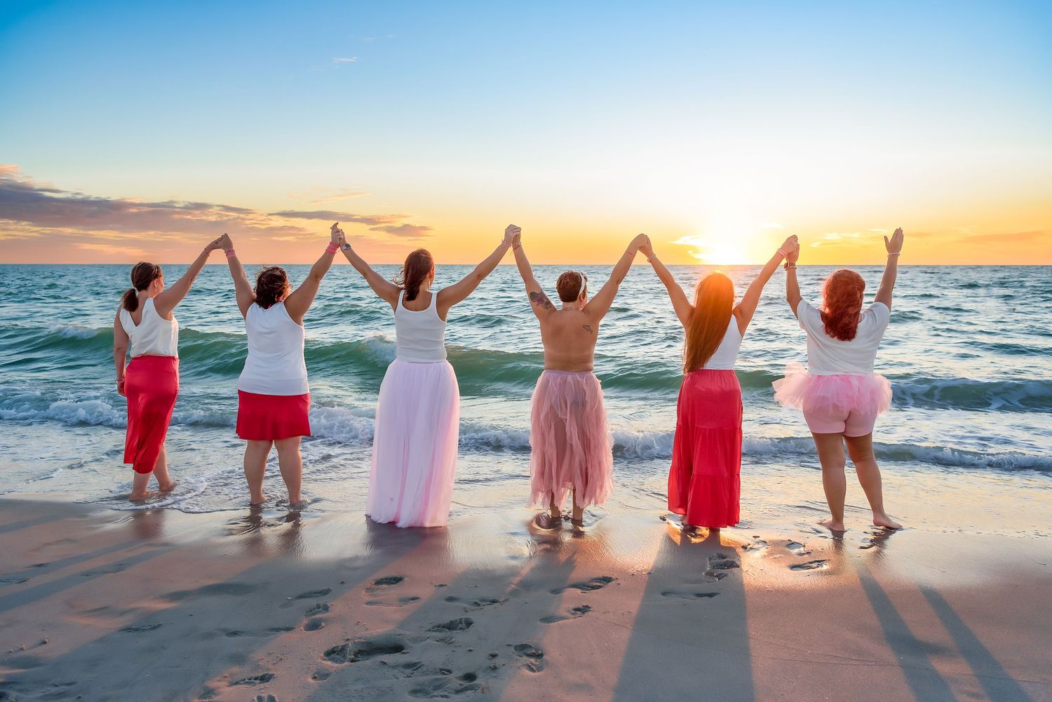 Group celebrating at sunset on a beach with raised arms against an orange sky.