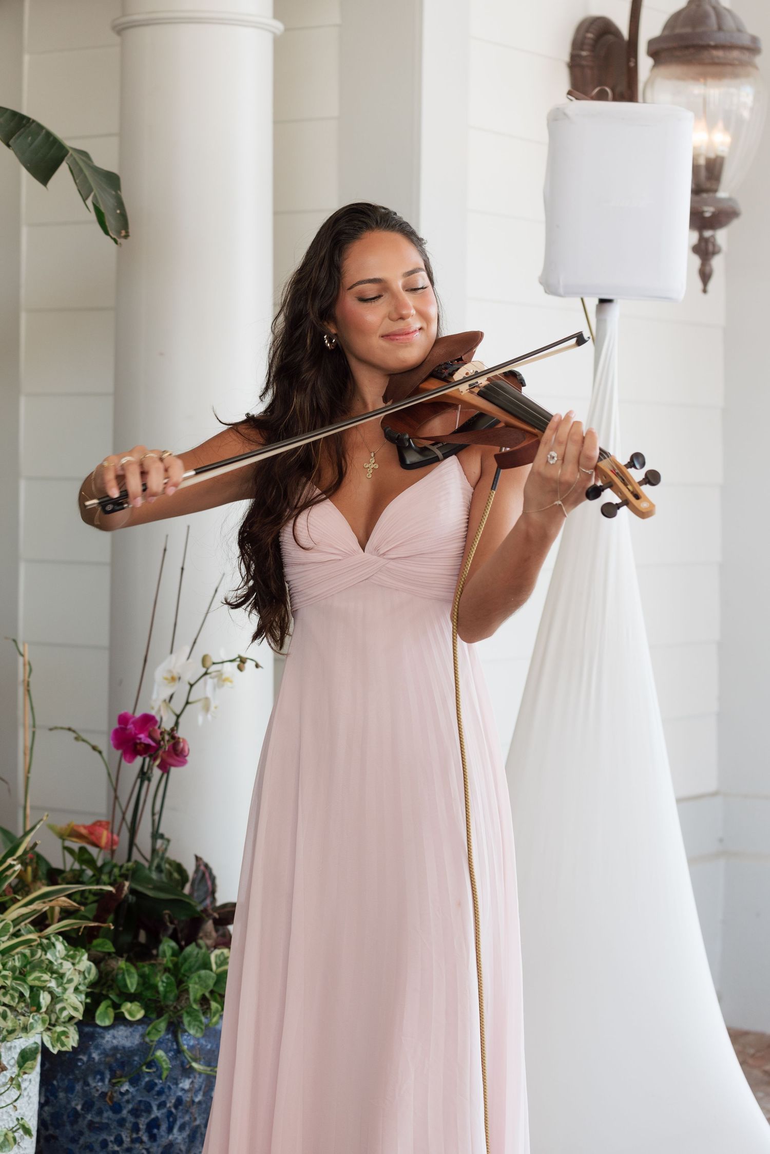A musician in a pink gown plays violin while standing by a white pillar with orchids and greenery.