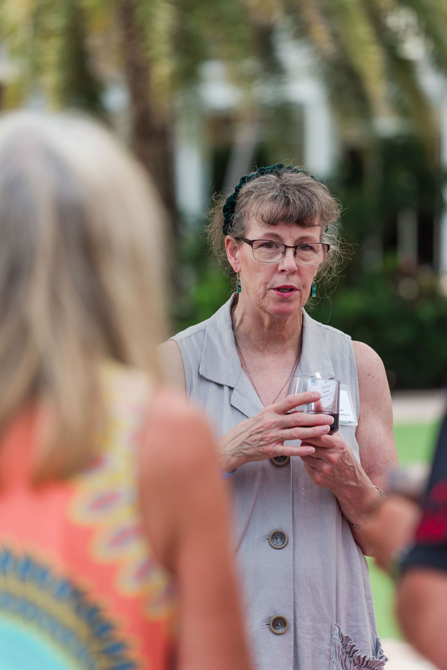 An older woman wearing glasses holds a drink while conversing outdoors near palm trees.
