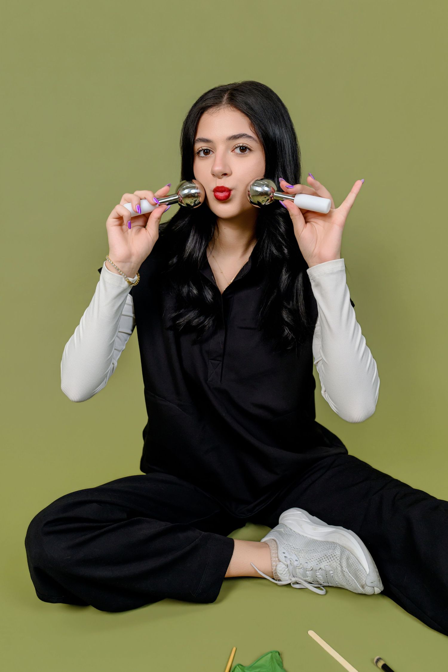Young woman in black outfit sitting cross-legged on green background, holding silver facial massage rollers near her face.