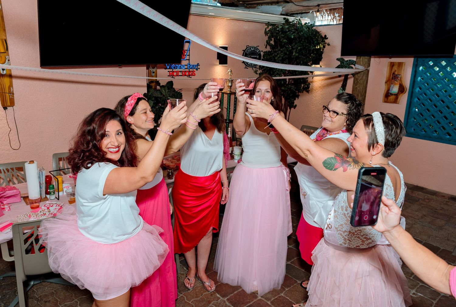 A group in pink tutus and party attire dance together with raised arms during a festive celebration.