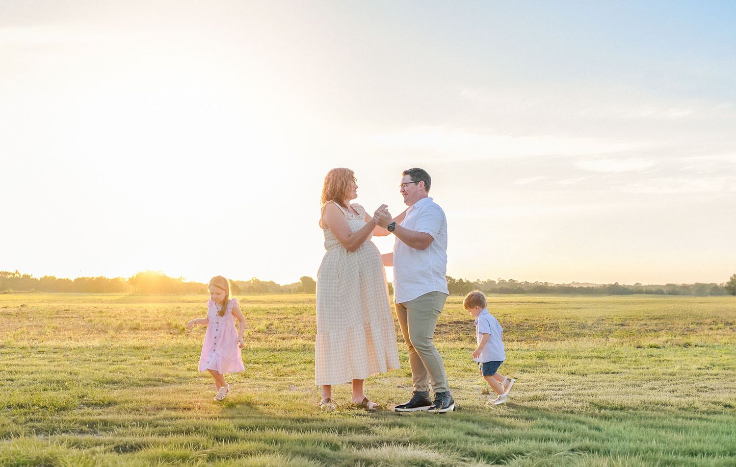 A family shares a tender moment together in a sunlit meadow during golden hour as two children play nearby.