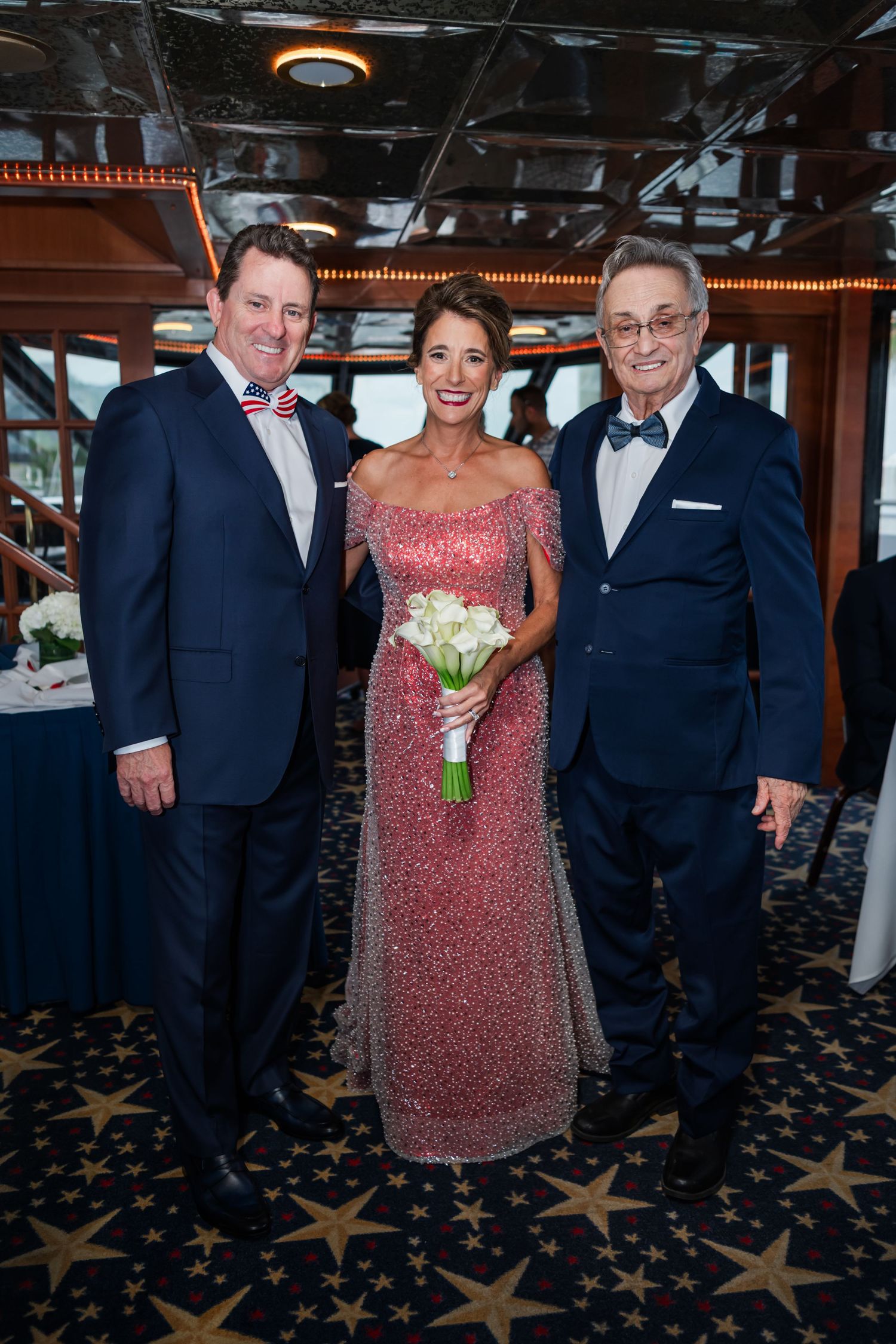 Three elegantly dressed guests pose together at a formal event, woman in pink beaded gown holding white bouquet.