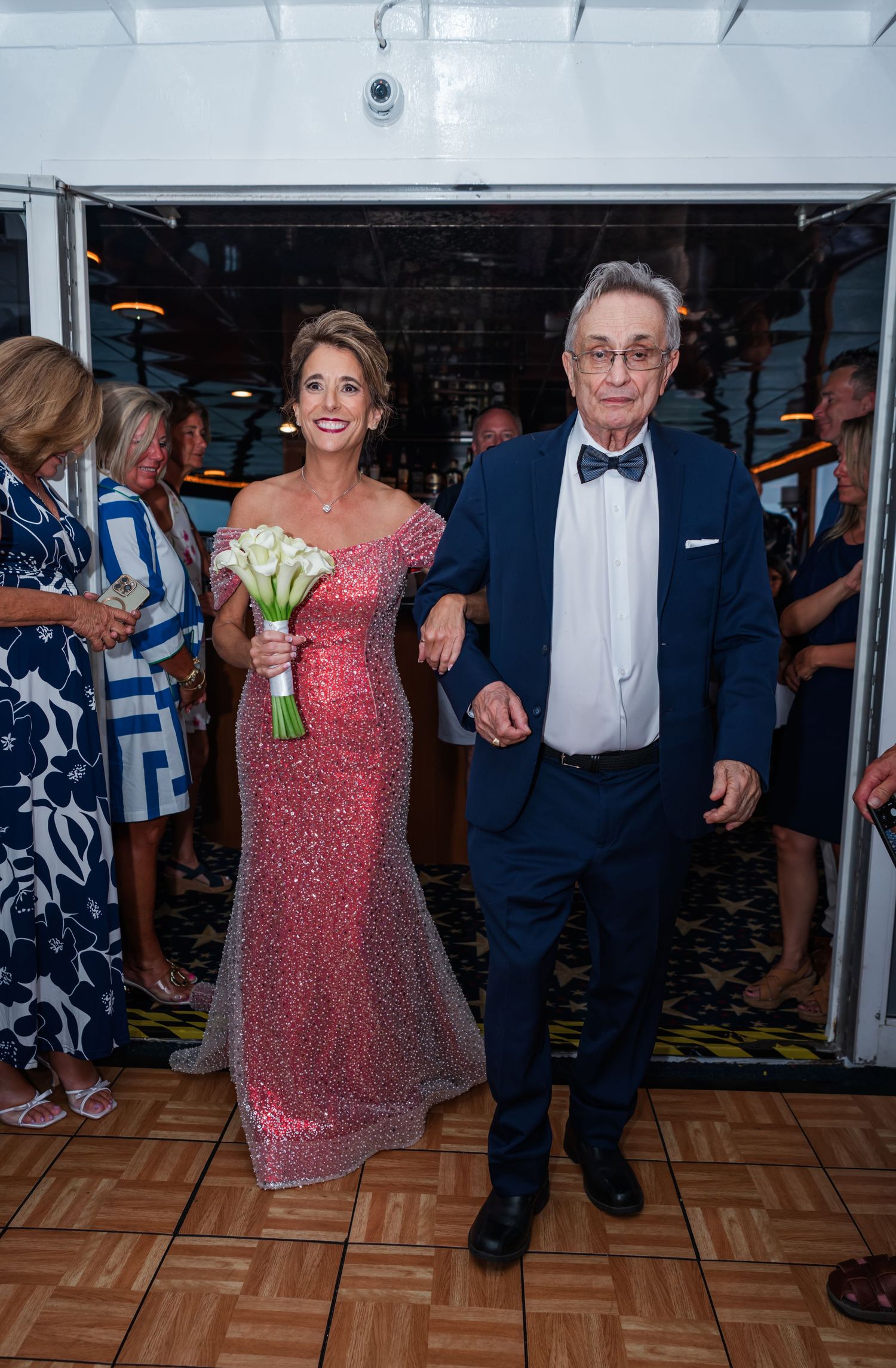 Elegant couple in formal attire walking together, woman in pink sequin gown holding white bouquet, man in navy tuxedo.