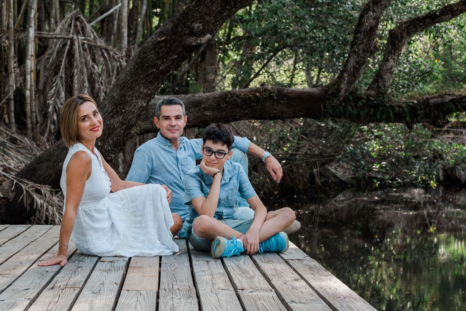 Family sits together on wooden dock by tranquil pond under overhanging tree branches on summer day.