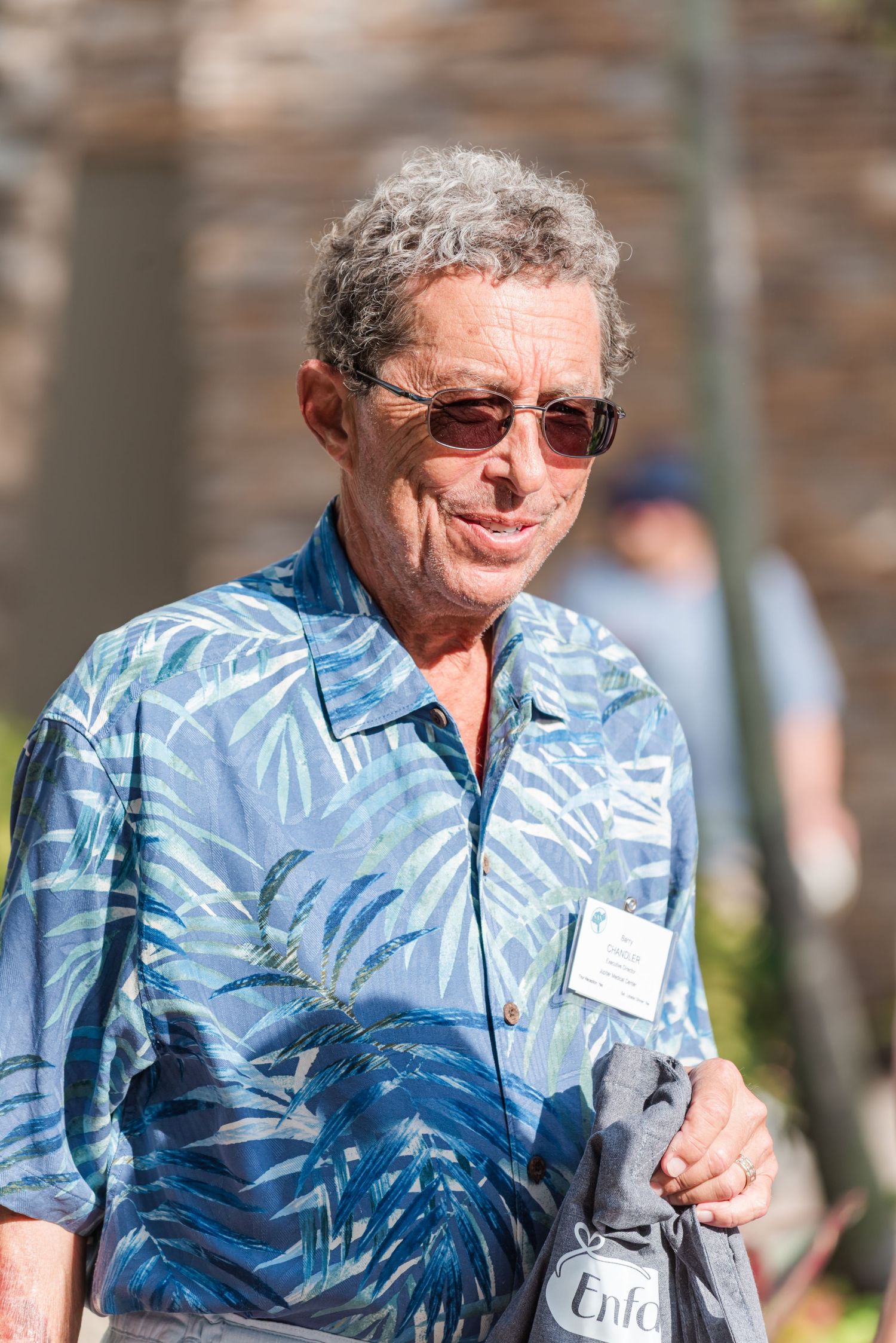 Smiling man in a blue tropical print shirt and sunglasses wearing a name tag at an outdoor event.