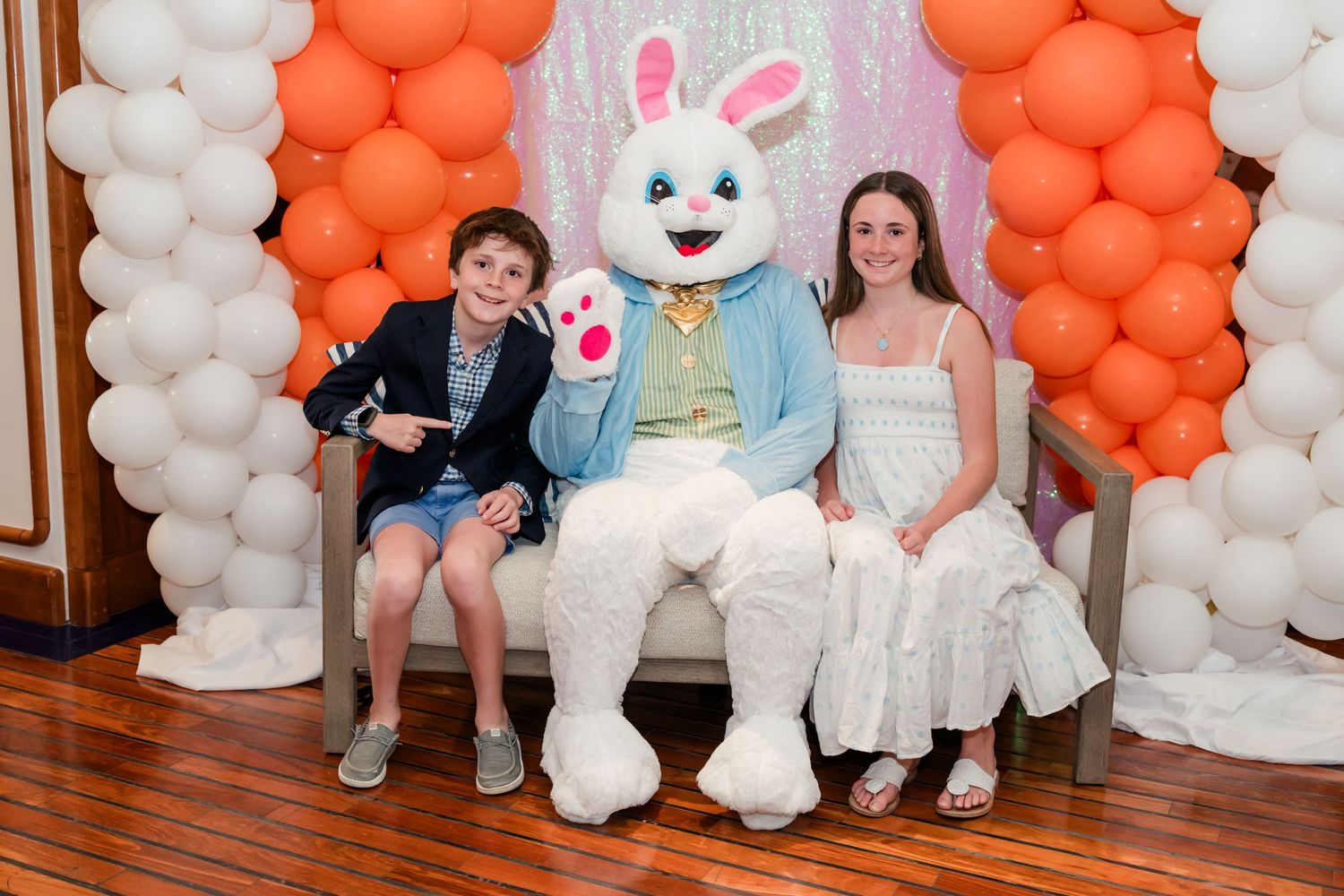 Two children posing with Easter Bunny mascot surrounded by orange and white balloon arch decorations.