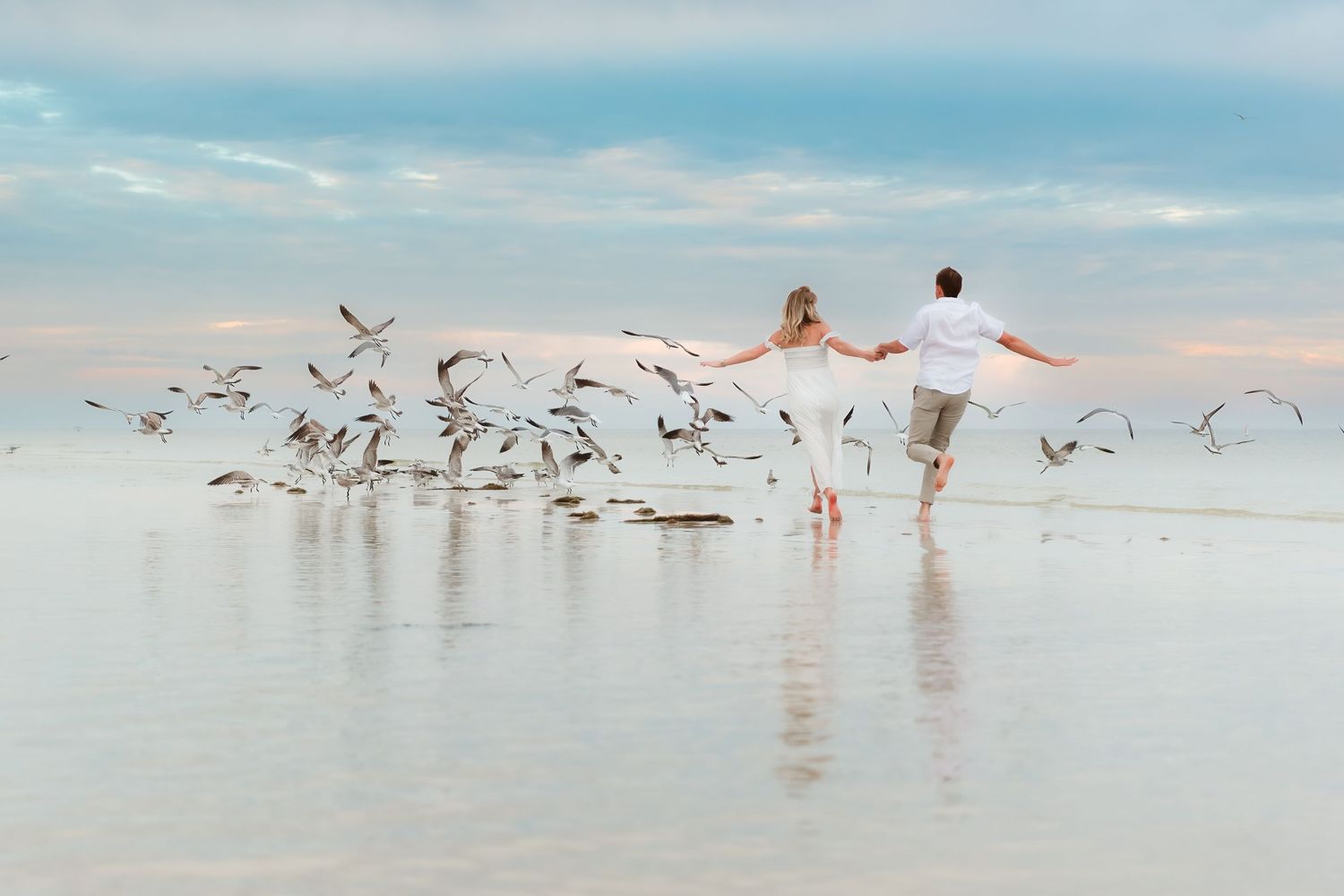 Couple running hand-in-hand on a beach at sunset, scattering a flock of seagulls over shallow reflective water.