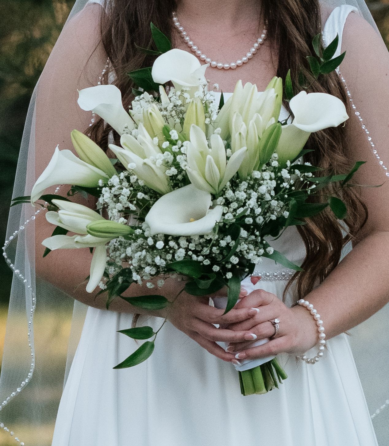 Bride in white gown holding elegant bouquet of white lilies, calla lilies, and baby's breath, wearing pearl jewelry.