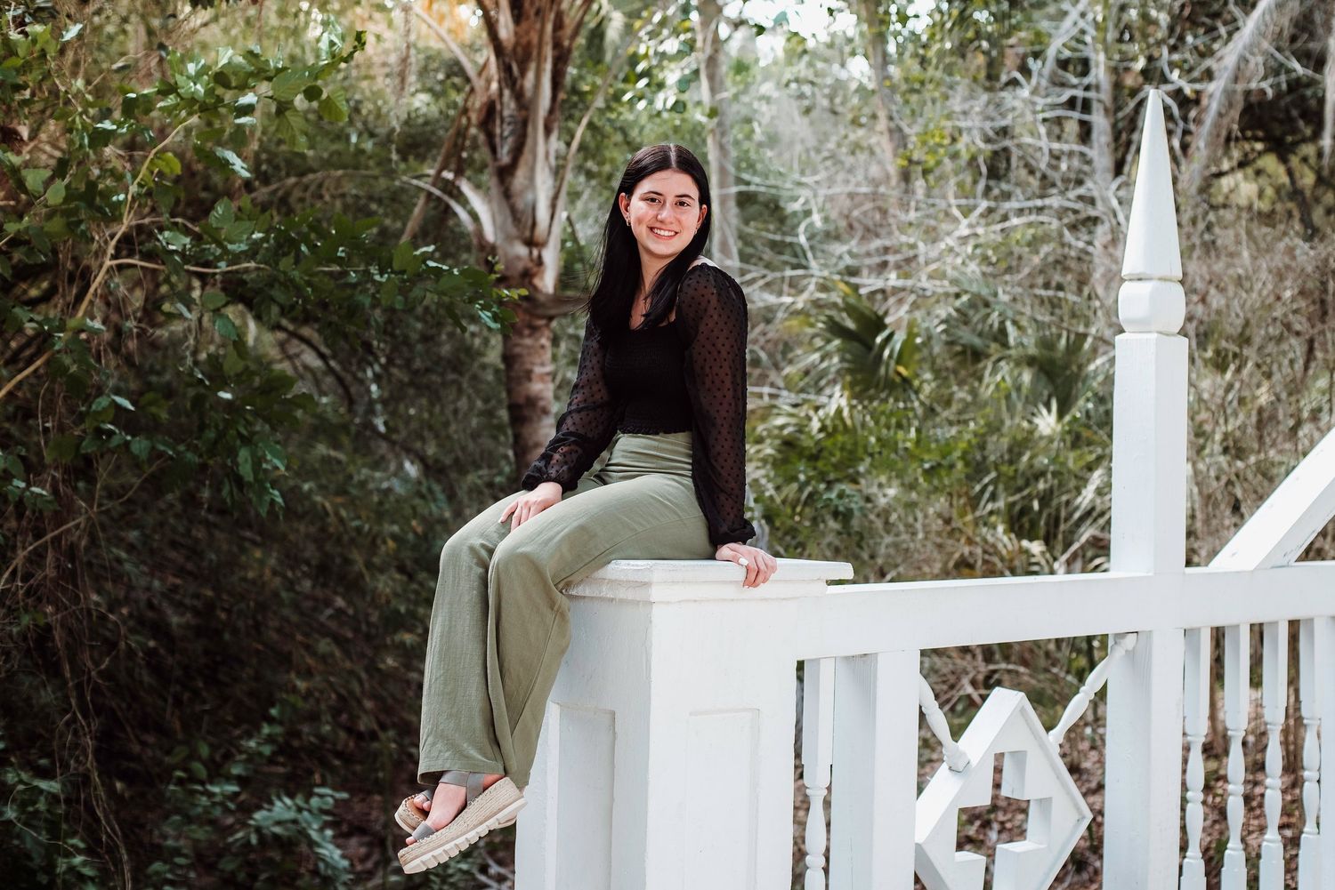 Person in a black top and light colored pants sits casually on white garden fence railing surrounded by greenery.
