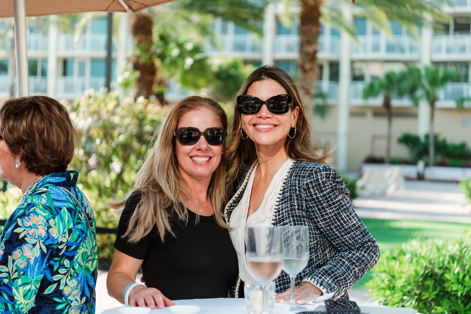Two people in sunglasses smile while enjoying wine outdoors at a tropical resort with palm trees in the background.