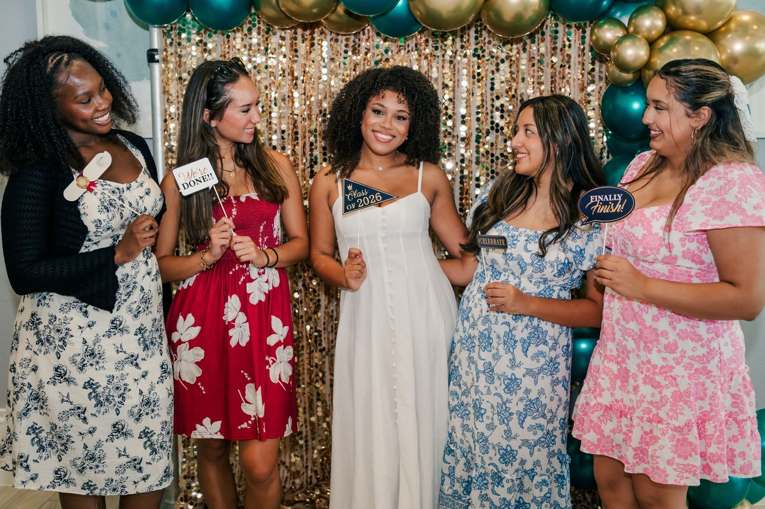 Five women in floral dresses laugh together at a party, holding photo booth props against a gold sequin backdrop.