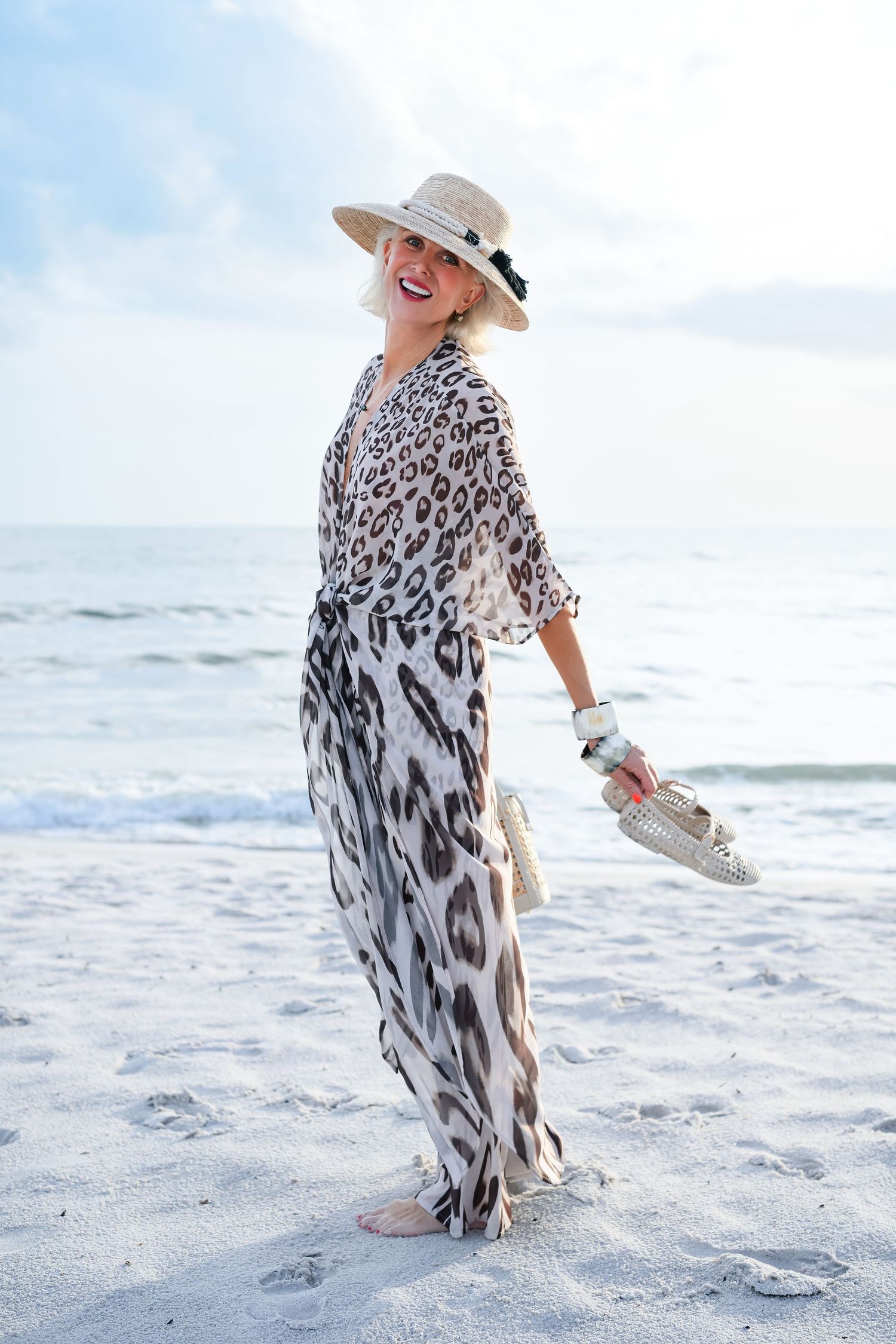 Woman in leopard print maxi dress and straw hat smiling on sandy beach holding shoes with ocean waves behind her.
