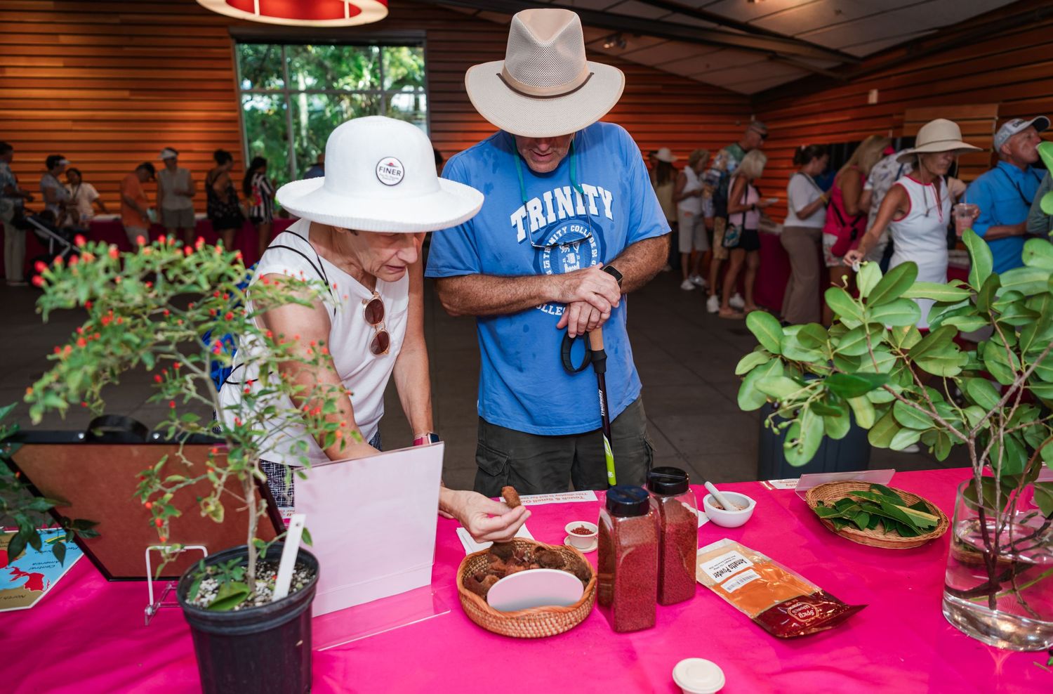 Two people in sun hats examine plants and products at a vibrant outdoor garden market event.