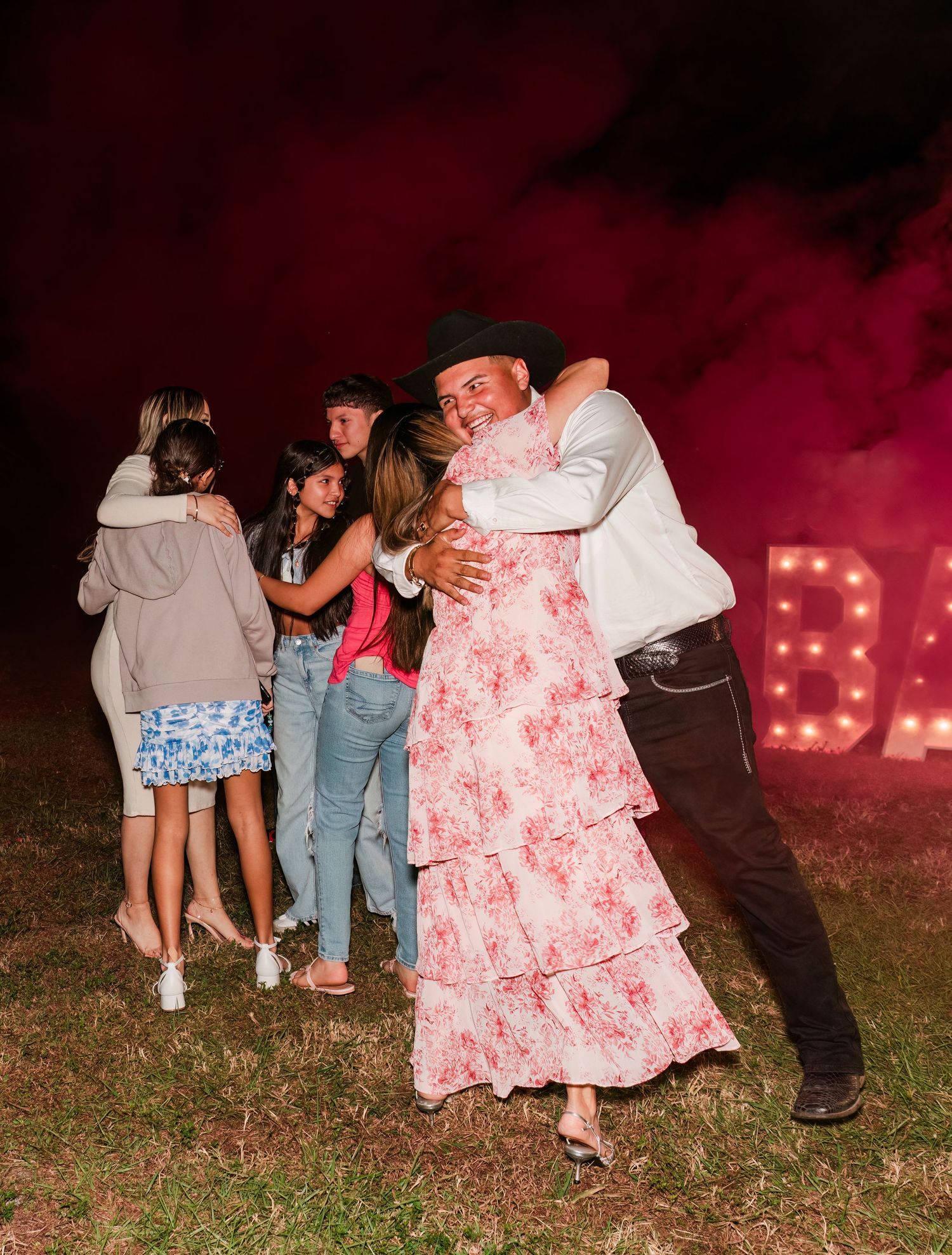 People dancing and celebrating at an outdoor party with red lighting and smoke effects in the background.