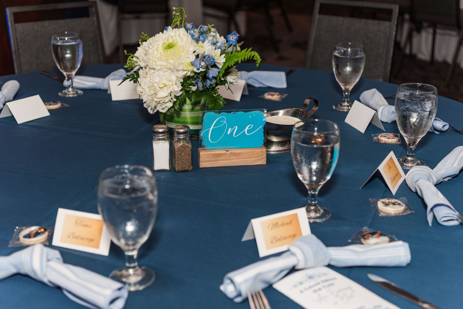 Wedding reception table setting with navy blue tablecloth, white floral centerpiece, and turquoise table number card.