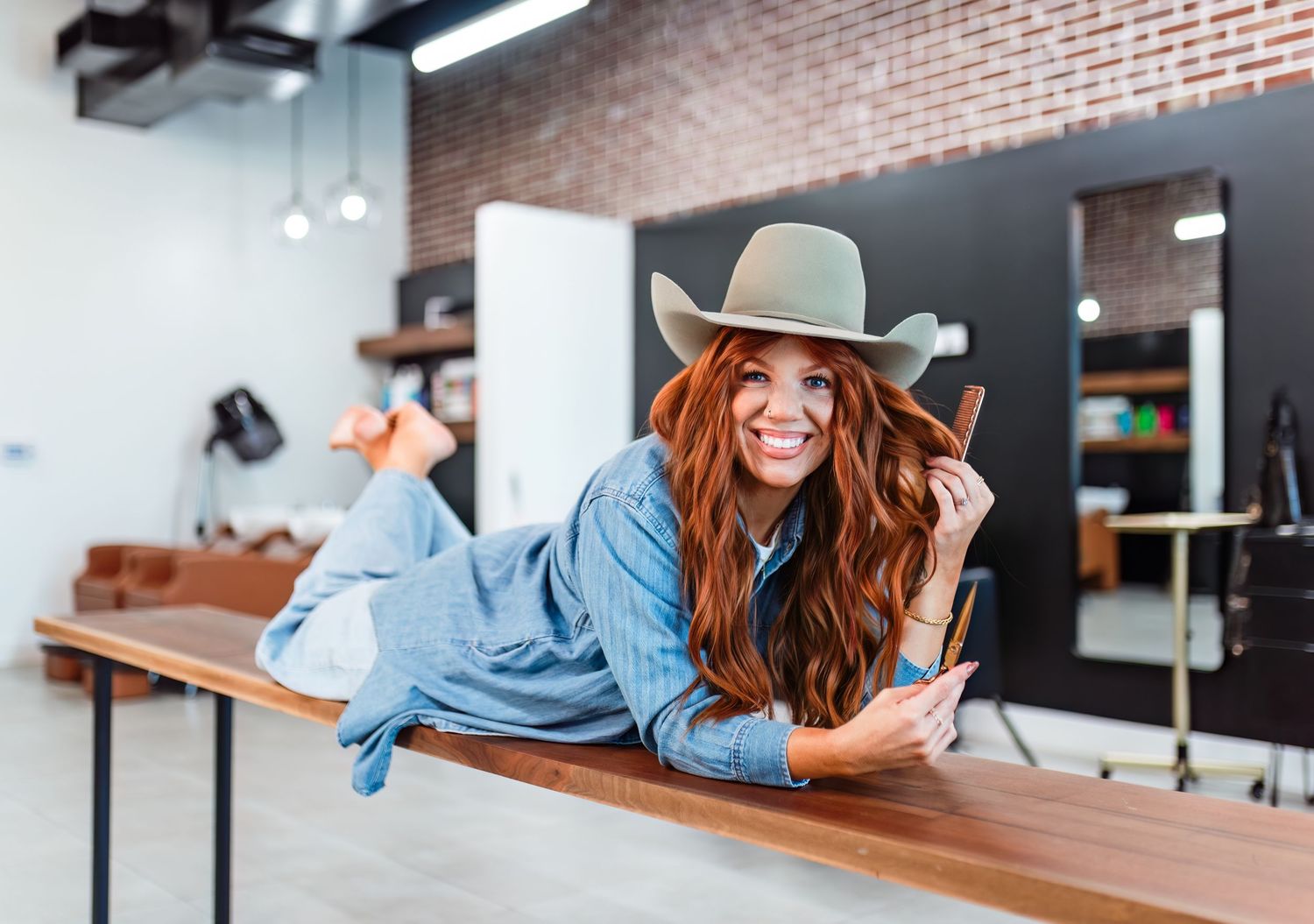 Woman in denim shirt and cowboy hat lies on wooden bench while smiling at camera in brick wall studio setting.