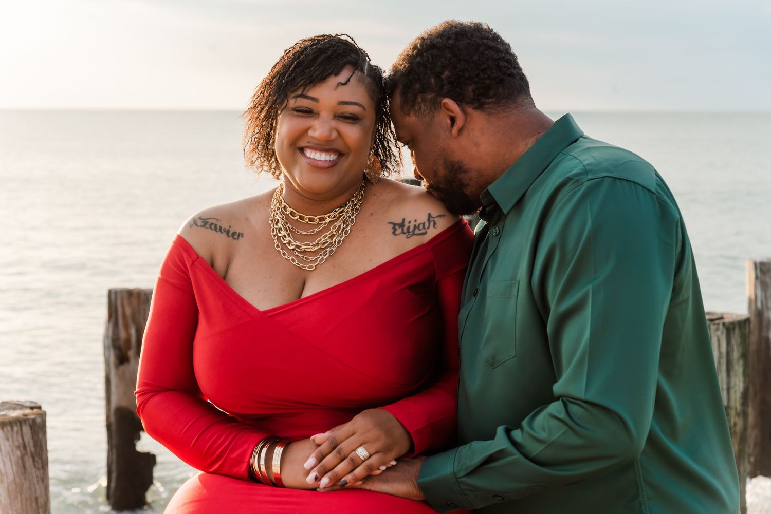 Couple shares intimate romantic moment together on beach pier at sunset while wearing red and green sweaters.