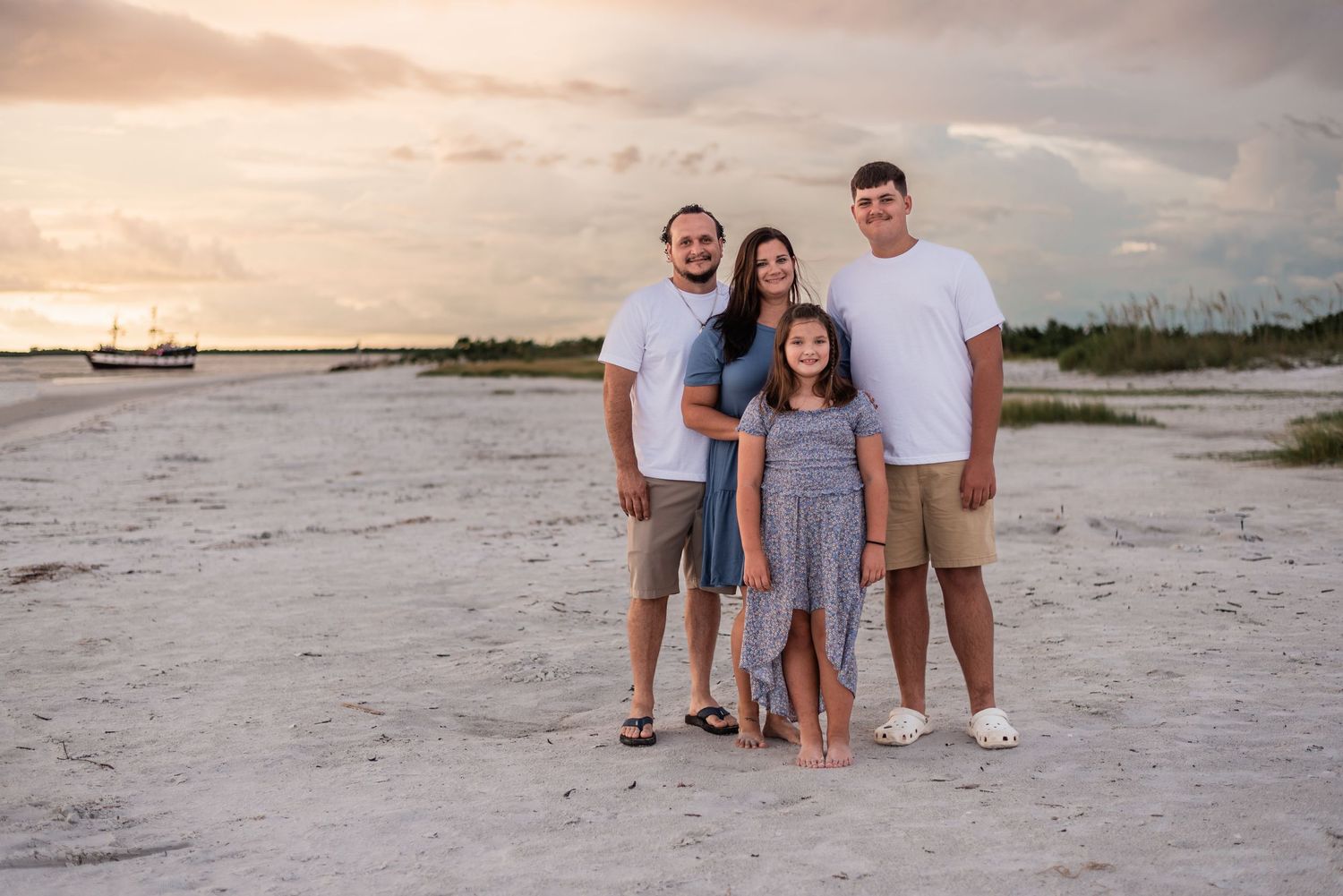 A group of four people wearing white and blue outfits pose together on a sandy beach at sunset.