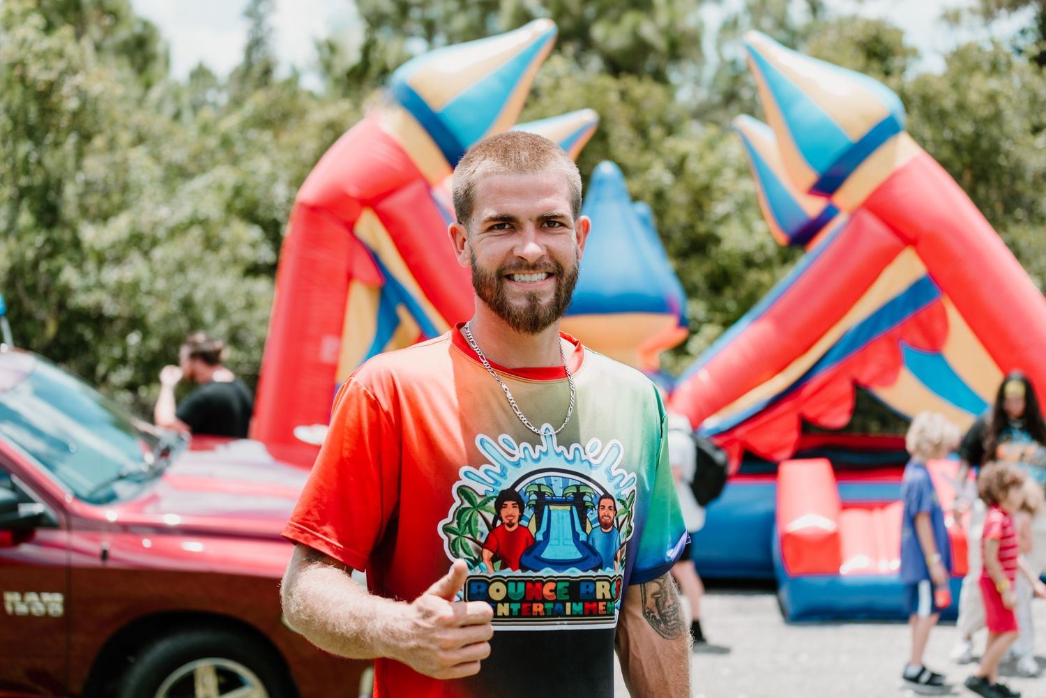 Person holding a colorful event t-shirt and smiling at outdoor festival with bounce houses in background.